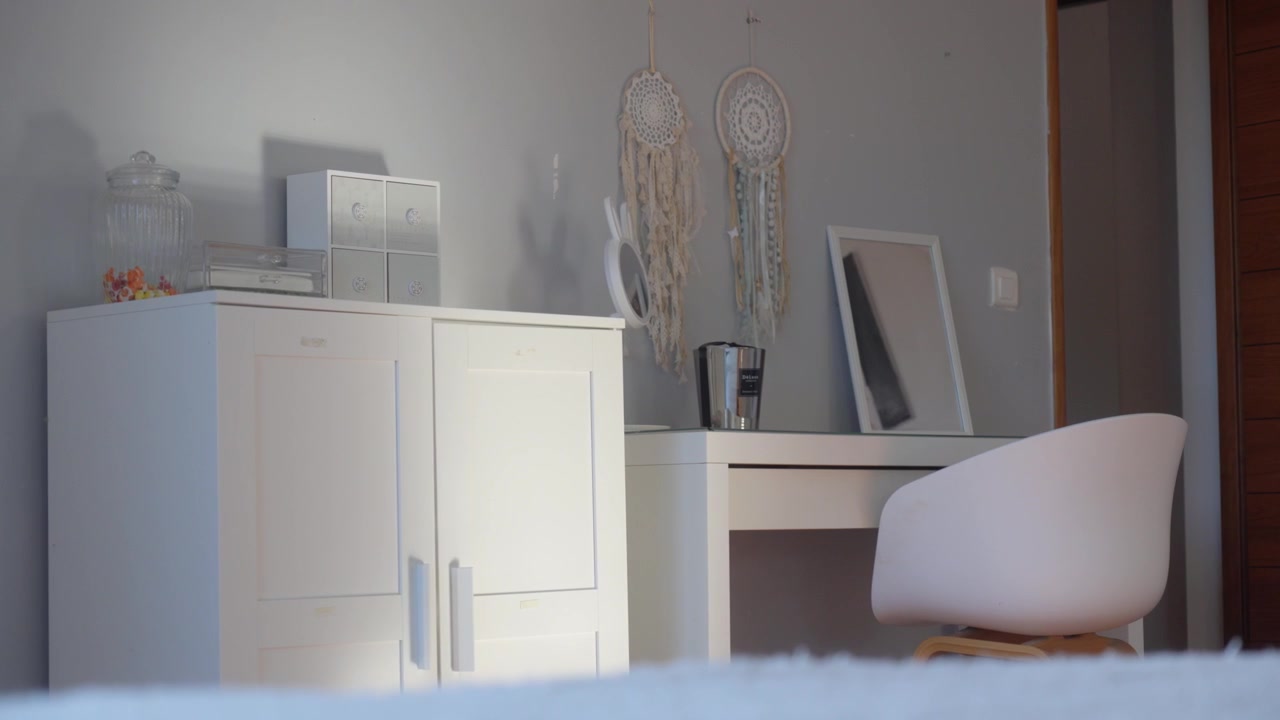Slow arc shot of a bright, cozy bedroom interior with white cabinets, a desk, chair, and boho dreamcatcher wall decor. Warm diffused natural light creates a calm, welcoming mood.
