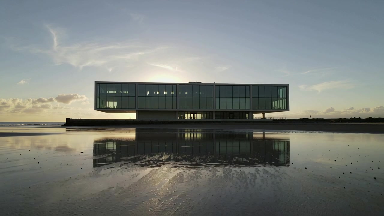Il sole che tramonta retroillumina un moderno edificio in vetro su una piattaforma rialzata in riva al mare, con la sabbia bagnata che riflette la luce