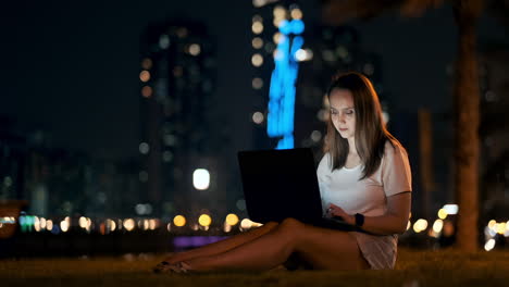 Eine-Junge-Frau-Sitzt-In-Einer-Sommernacht-Auf-Dem-Rasen-In-Der-Stadt,-Schaut-Auf-Den-Laptop-Bildschirm-Und-Schreibt-Eine-Nachricht-Auf-Die-Tastatur.-Freiberufliche-Programmiererin.-Nachtarbeit-Aus-Der-Ferne