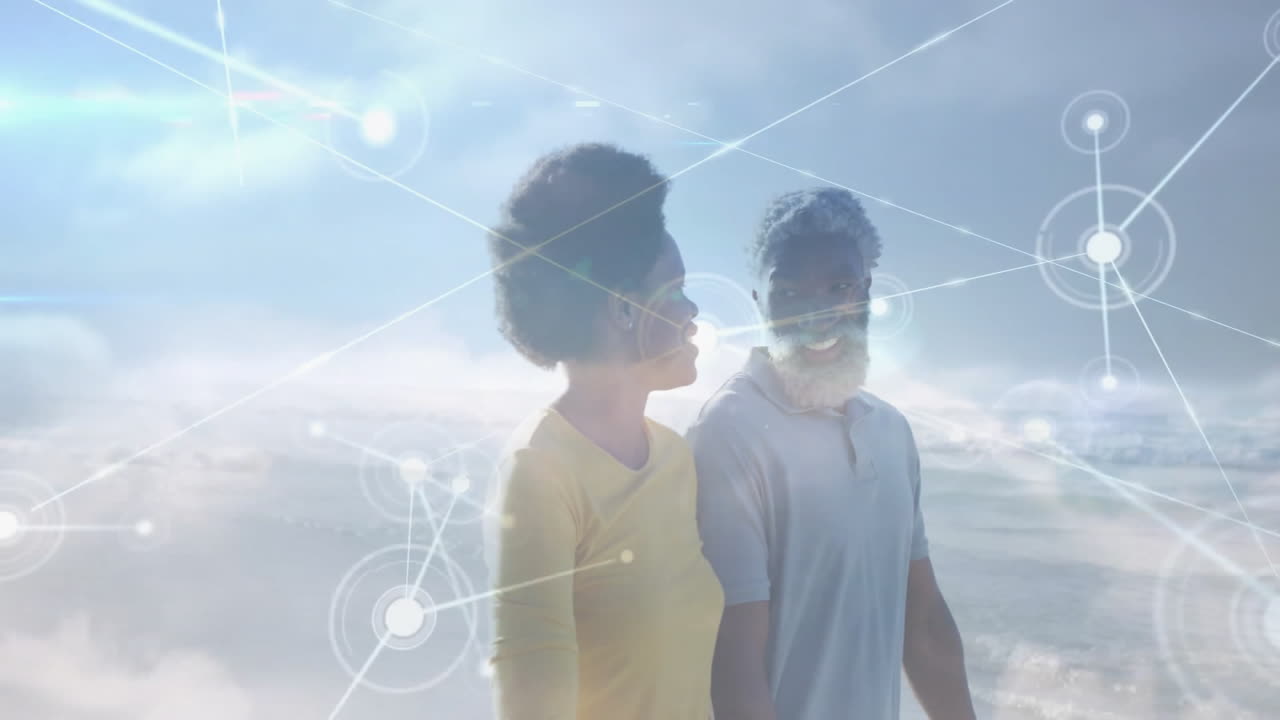 Mid adult woman and mature man walking beach shoreline, showing digital technology network overlay