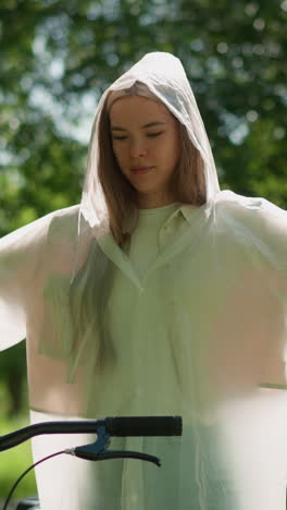 una mujer joven con un impermeable translúcido se para con los brazos extendidos, balanceando suavemente sus manos cerca de su bicicleta en un día soleado, rodeada de exuberante vegetación