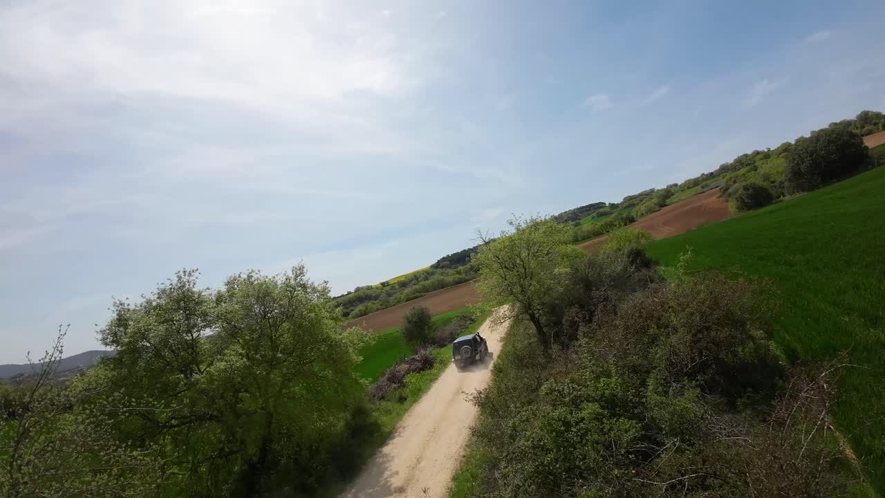 A drone hovering over a green field at low altitude. A 4x4 off-road vehicle appears from left to right, and the drone follows the vehicle from above along the dirt road. Aerial pursuit