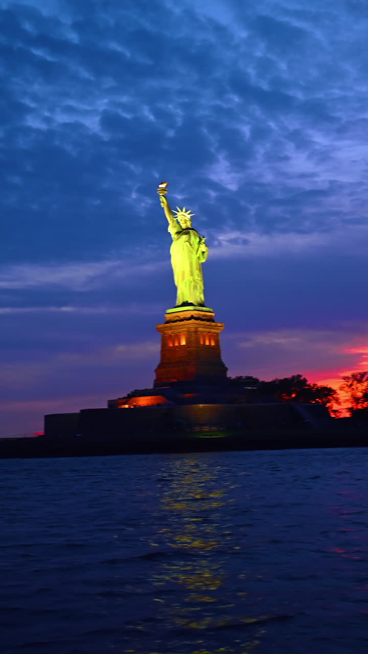 New York, AS, 8 Agustus 2025: Patung Liberty diterangi cahaya terang. Sudut pandang rendah pada landmark dengan latar belakang langit mendung dari pemandangan sungai. New York, AS. Video vertikal