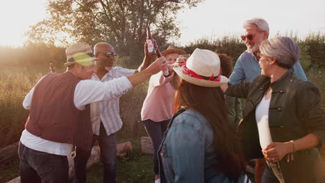 Eine-Gruppe-Reifer-Freunde-Macht-Toast,-Während-Sie-Am-Feuer-Sitzen-Und-Lieder-Auf-Dem-Campingplatz-Im-Freien-Singen