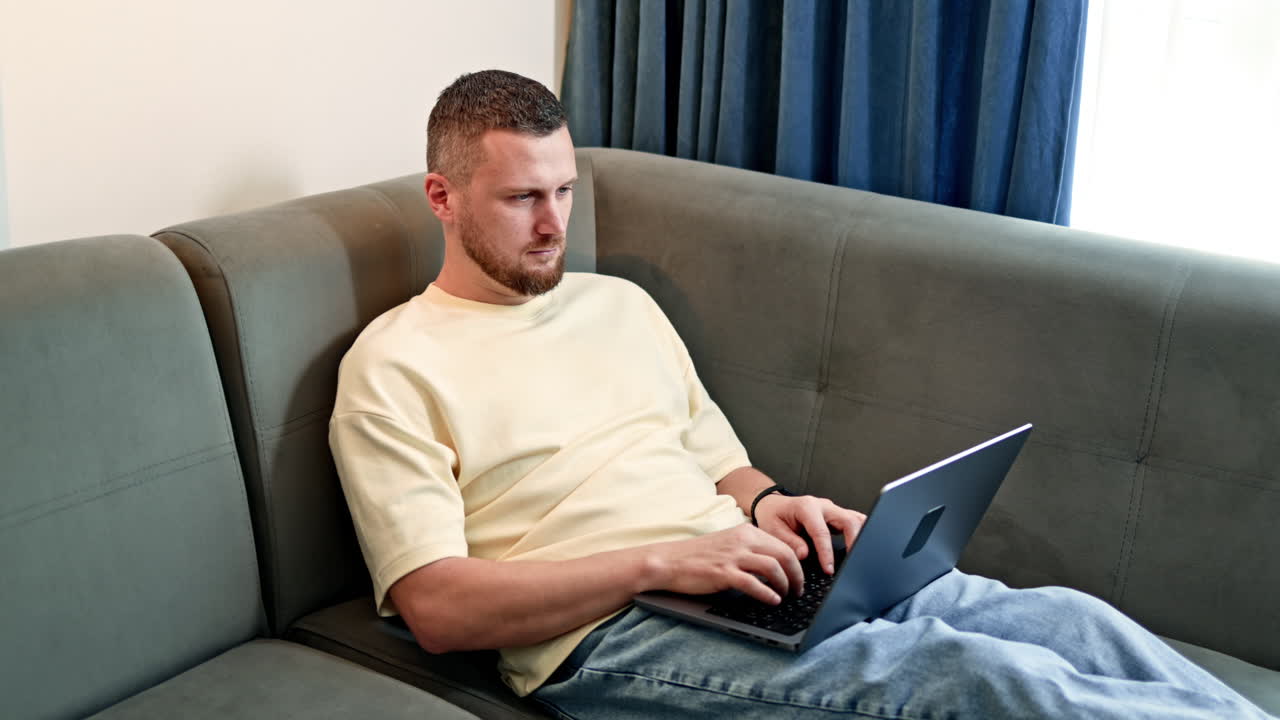 Mand arbejder på bærbar computer, mens han sidder i sofaen i stuen om dagen, fokuseret på skærmen med hænderne på tastaturet.