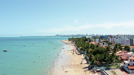 rising aerial wide shot of bessa beach in the tropical capital joao pessoa, brazil in the state of paraiba with brazilians and tourists enjoying the ocean and fishing boats at shore on a summer day