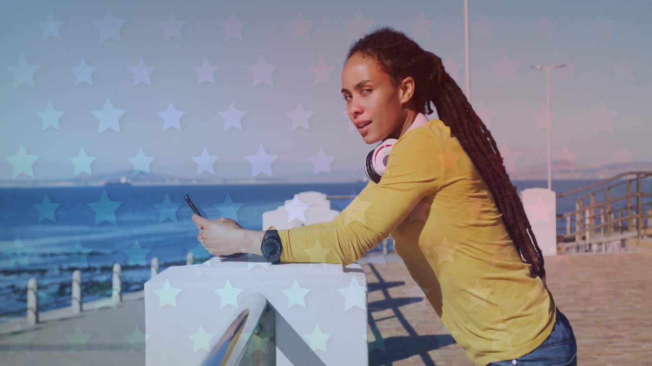 múltiples iconos de estrellas contra una mujer afroamericana que usa un teléfono inteligente en el paseo marítimo