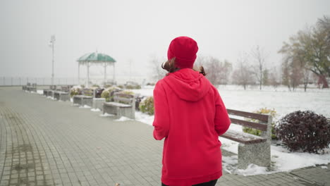 back view of woman jogging in red beanie and jacket along snowy pathway with iron railing on the left, serene winter park setting, benches nearby, foggy atmosphere with winter trees around