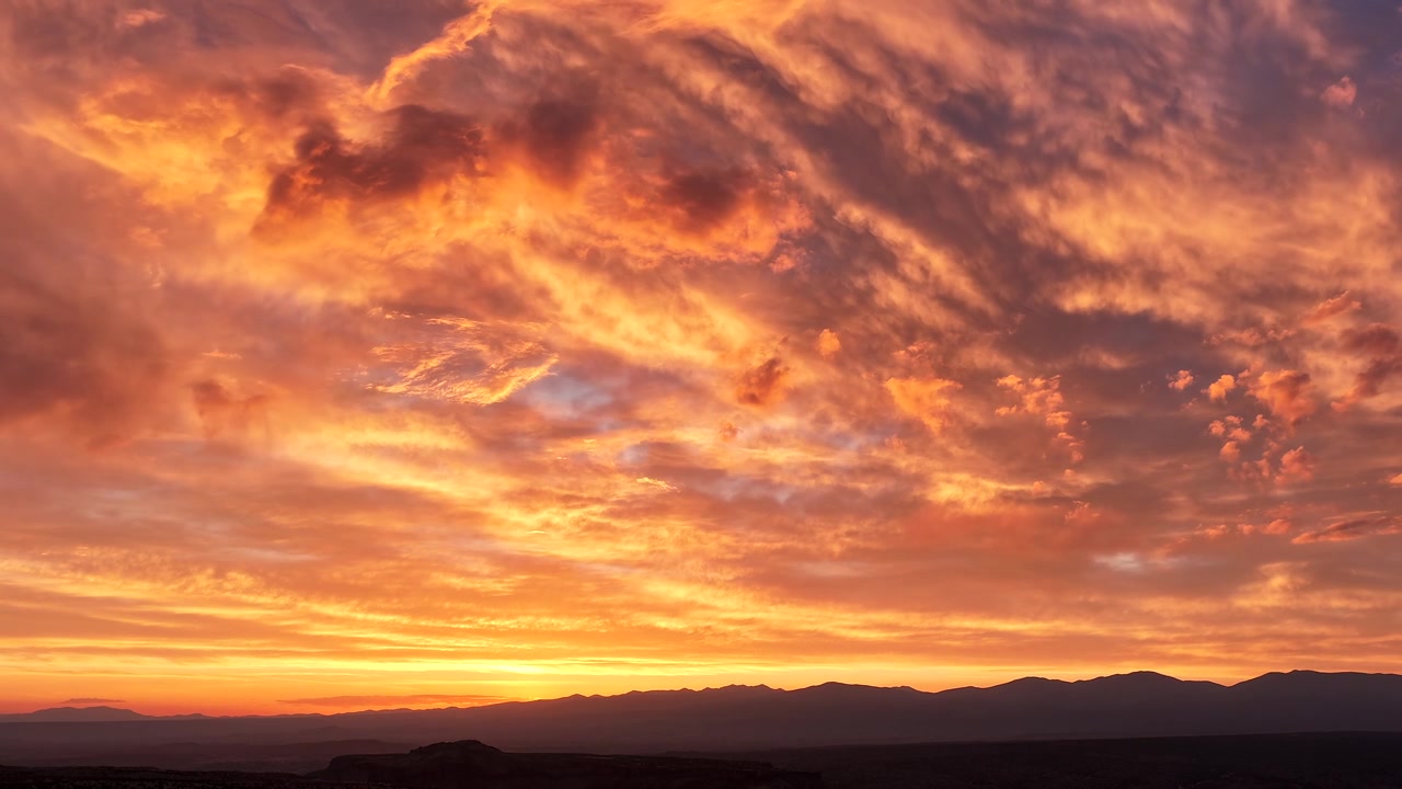 Timelapse 4K panorámico con dron de un amanecer brillante y ardiente que ilumina el cielo en vibrantes tonos rojos, dorados y naranjas. Nubes matutinas dramáticas evolucionan sobre la majestuosa silueta de una montaña oscura.