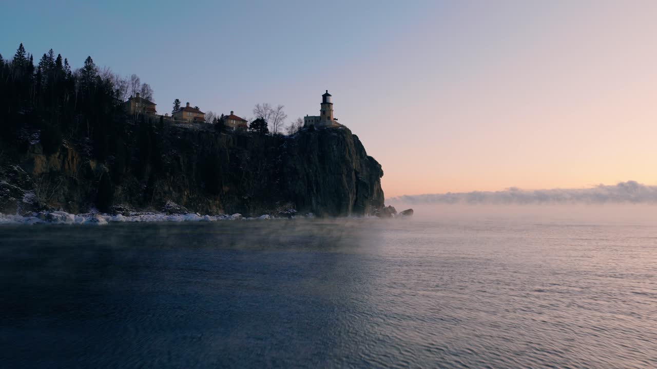 Una ripresa aerea stabile a bassa quota che si muove sulle acque increspate del Lago Superiore verso il faro di Split Rock durante un'alba invernale nebbiosa.