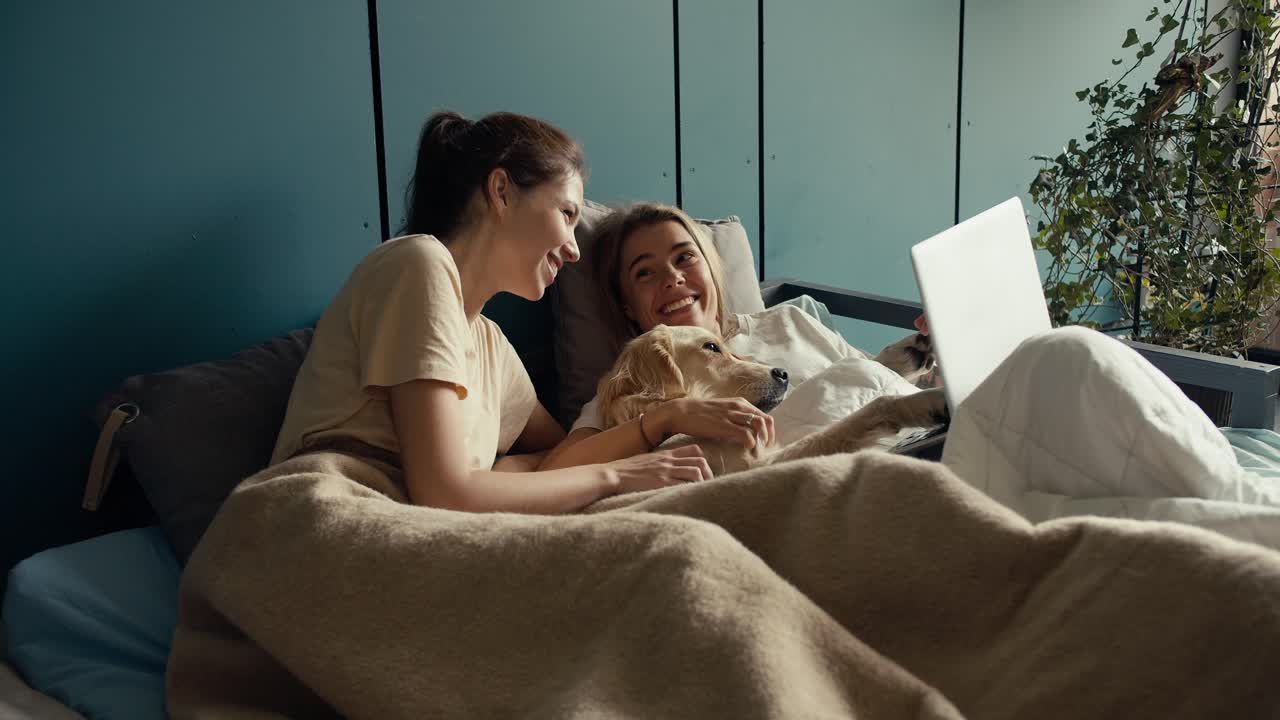 금발 소녀와 갈색 소녀가 노트북 스크린을 보고 얘기하면서 밝은 색의 개와 함께 담요로 싸여 침대에 누워 있습니다.
