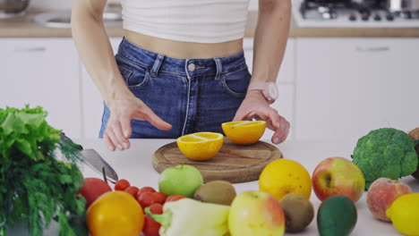 woman with sharp knife cutting orange into two halves. table full of fresh fruit and vegetables in the kitchen. woman preparing healthy food for losing weight. vertical video