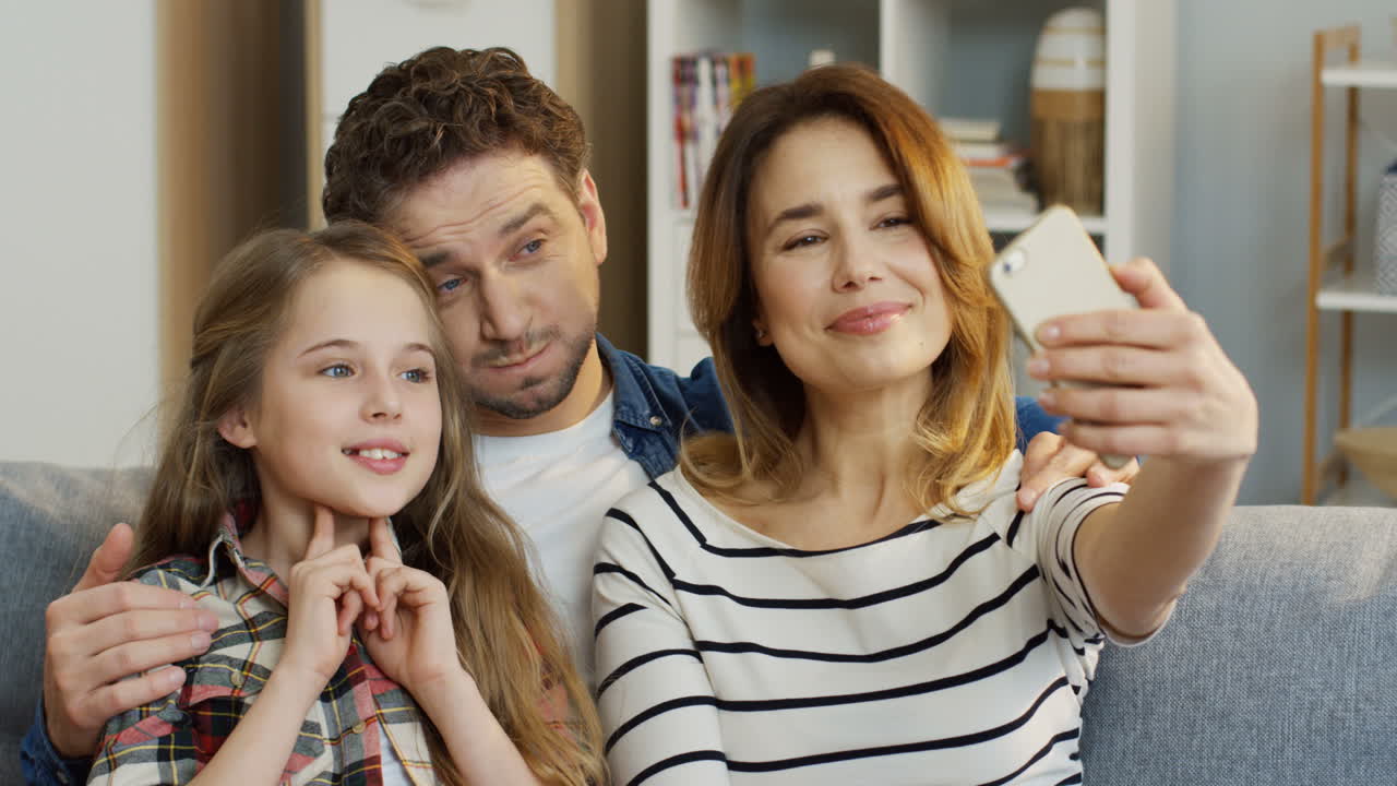 родители и дочь, сидя дома на диване, делают забавные селфи на смартфон, улыбаясь и позируя на камеру