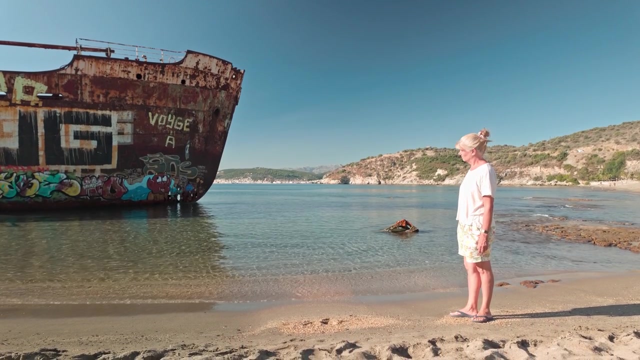 Frau betrachtet rostiges Schiffswrack, das an einem mediterranen Sandstrand verrottet.