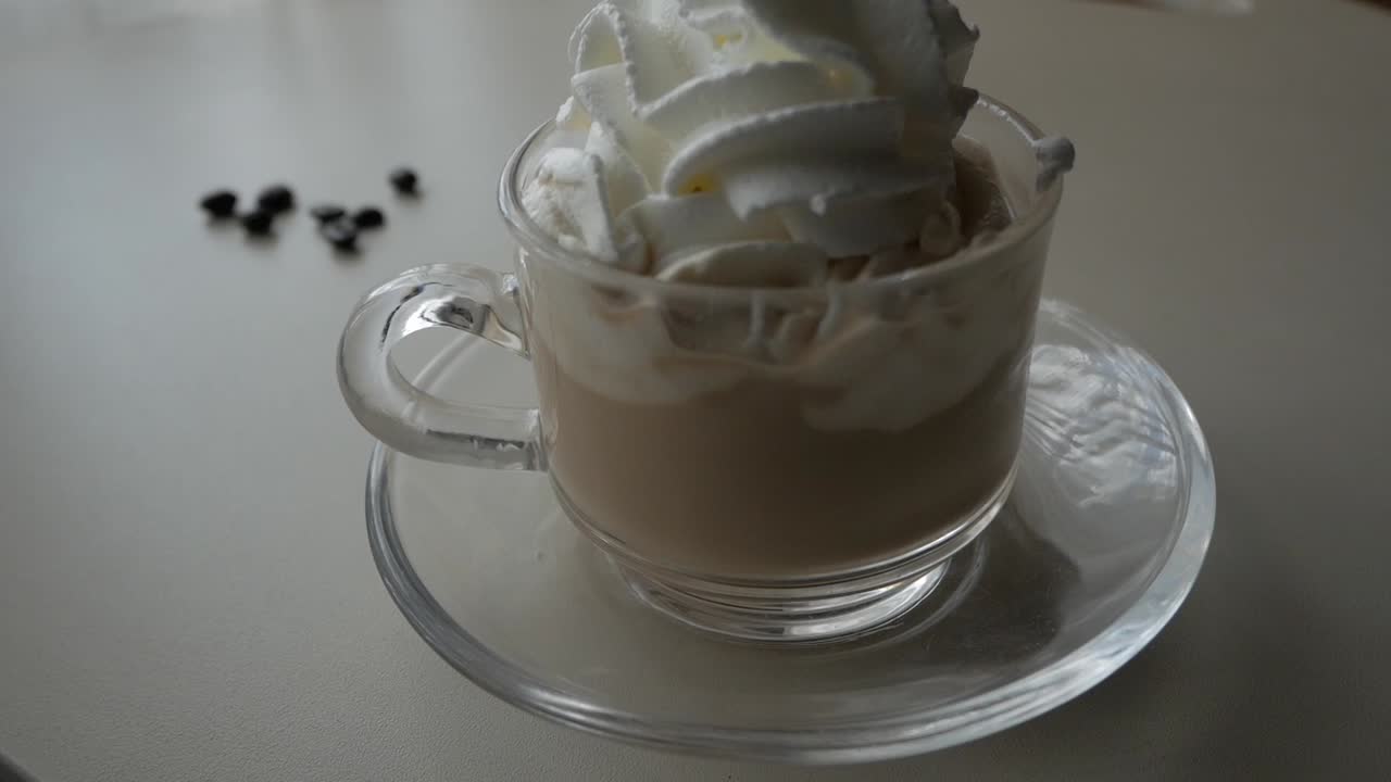 Side view of glass mug with coffee as whipped cream is poured inside, creating texture and foam under natural daylight