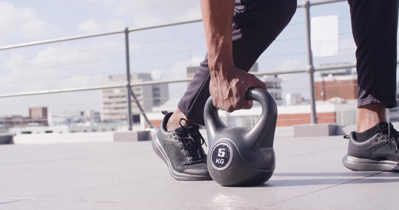 Uomo afroamericano con scarpe che cammina verso un kettlebell da 5 kg su un tetto, piegandosi e afferrandolo per l'allenamento.