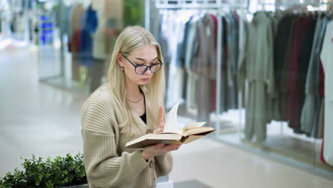lady in google glasses focuses on book in hand as she turns to the next page with green decorative flower behind, clothes well arranged seen through shop glass