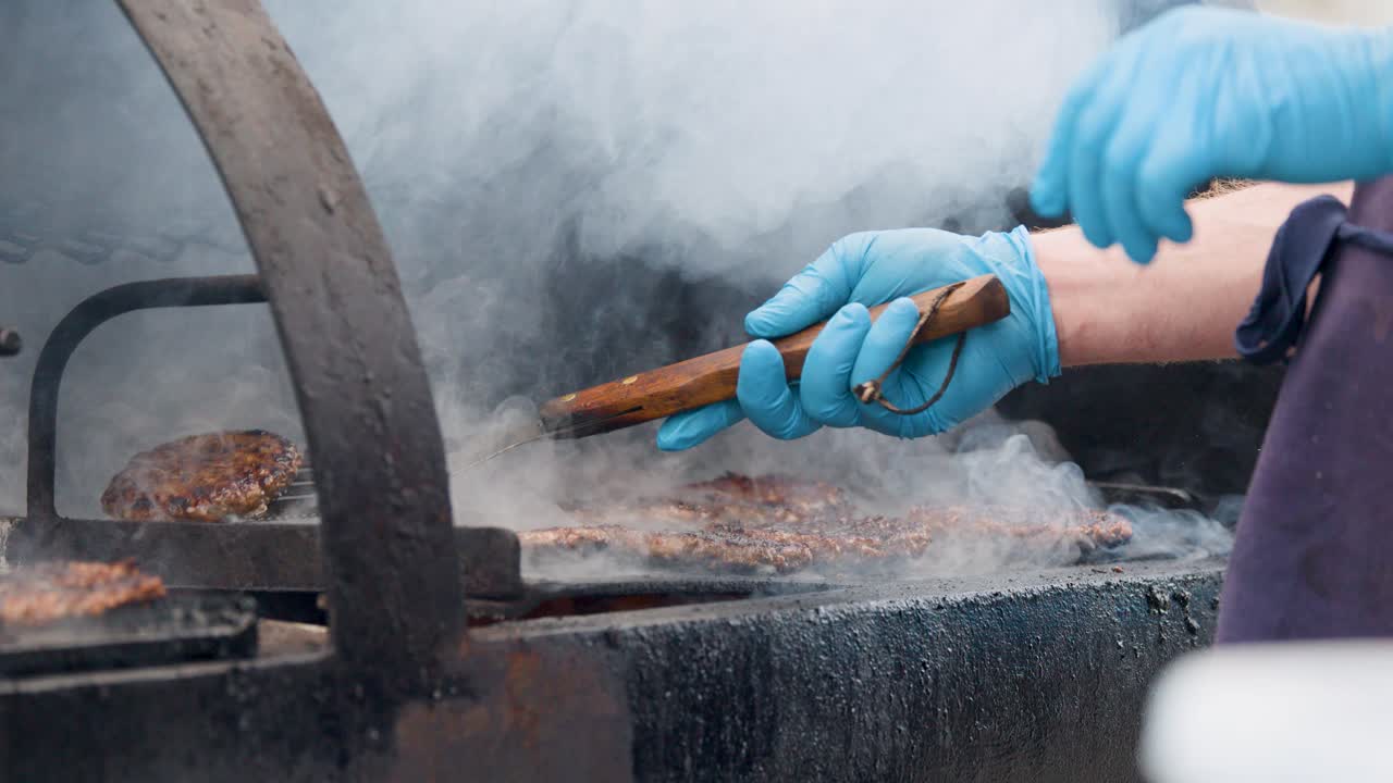 Person in blue gloves flips burgers on smoky grill using spatula, outdoor daylight, close-up