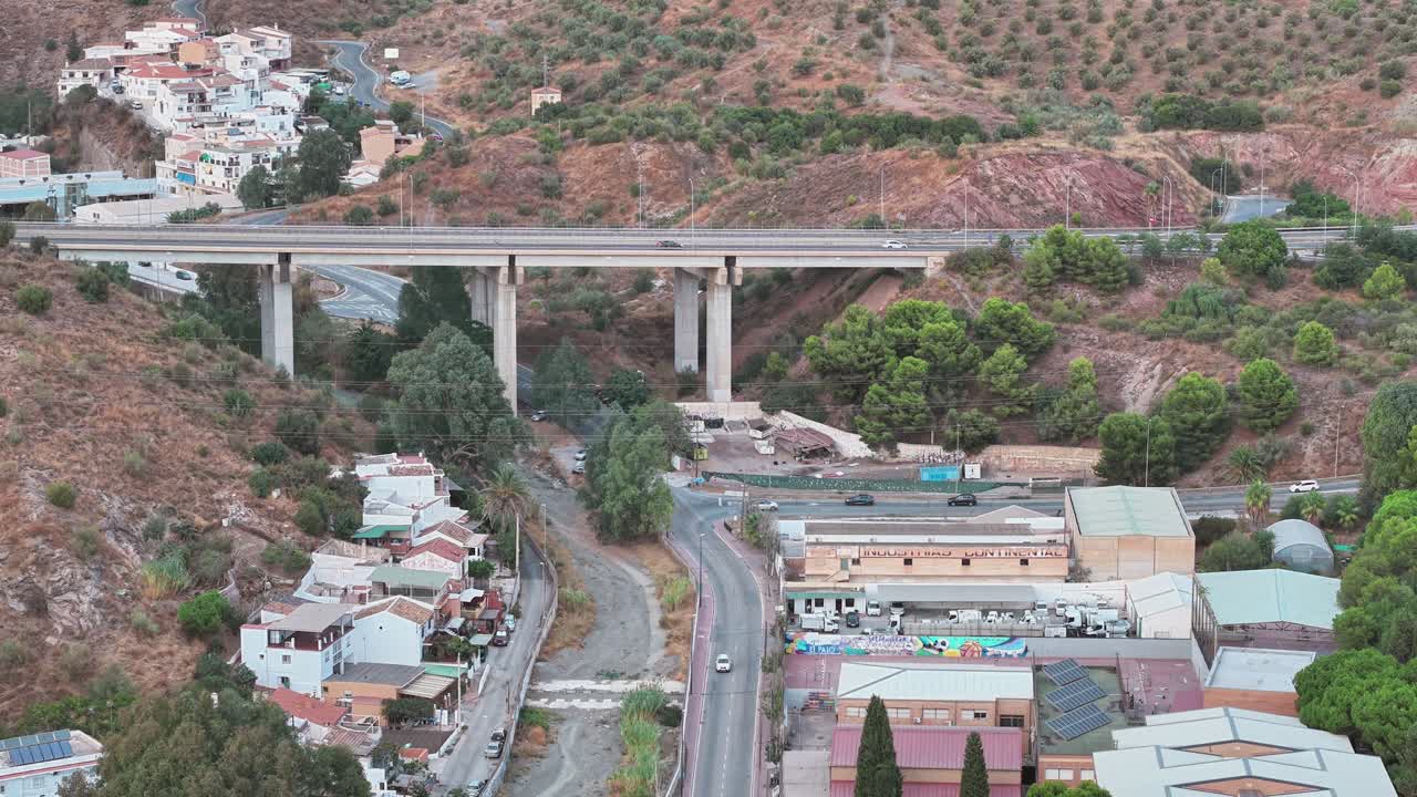 Aerial view of the outskirts of Málaga showing a scenic bridge spanning a valley, surrounded by hills, houses, and winding roads blending into the arid Mediterranean landscape