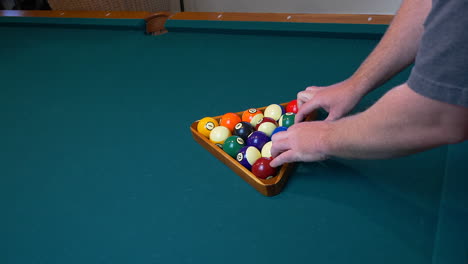 a caucasion man racking a set of billiard balls, for a game of eight ball, on a green felt pool table