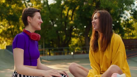 a brunette girl in a white sweater and a girl with a short haircut in a purple top and red headphones communicate while sitting on the floor in a skate park in summer