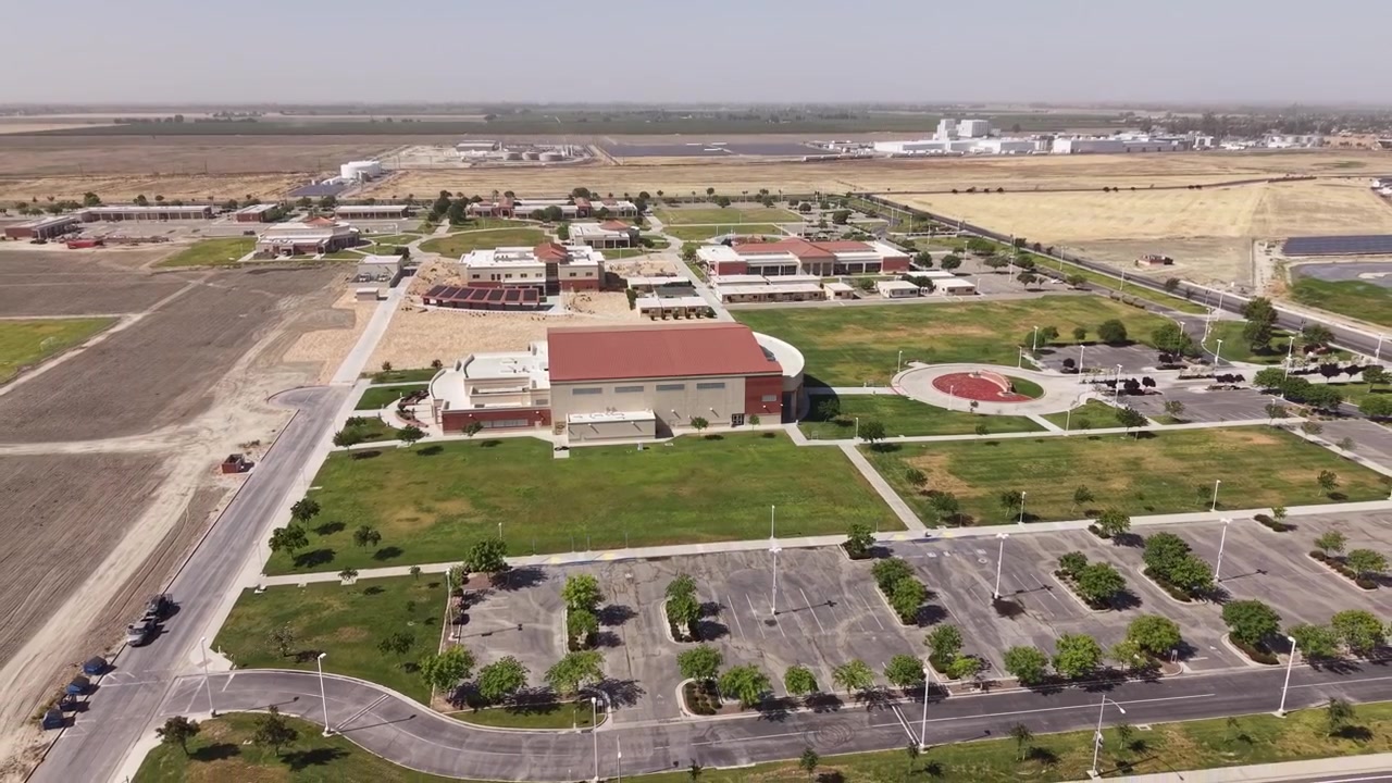Zoom indietro di drone dal campus del West Hills College Lemoore in una giornata di sole nella Central Valley della California. Mostra moderni edifici universitari, prati verdi e i terreni agricoli circostanti.