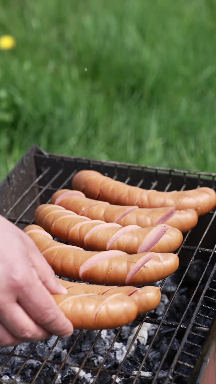 Man prepares juicy sausages in barbecue on picnic. Cook putting sausages into a grill for frying. Fried sausages are cooking on coals in the garden. Vertical video