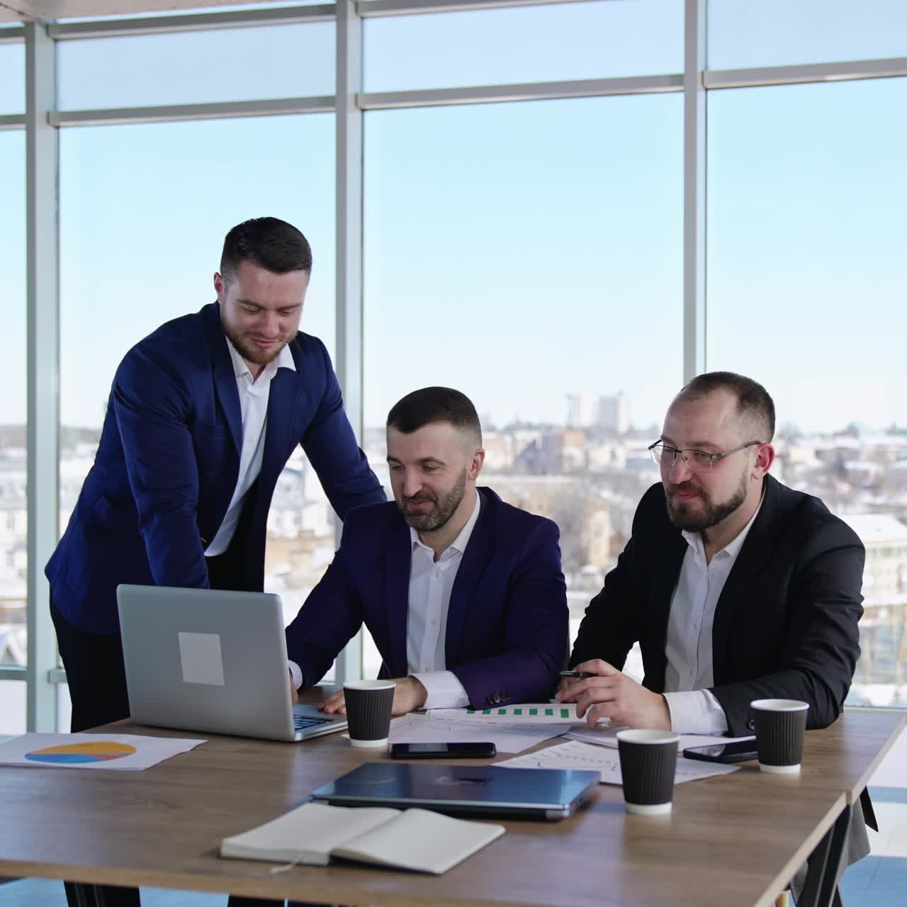 Three man near the desk discuss something and laugh. Two women look at the smartphone and talk about it. Panoramic windows with cityscape at backdrop