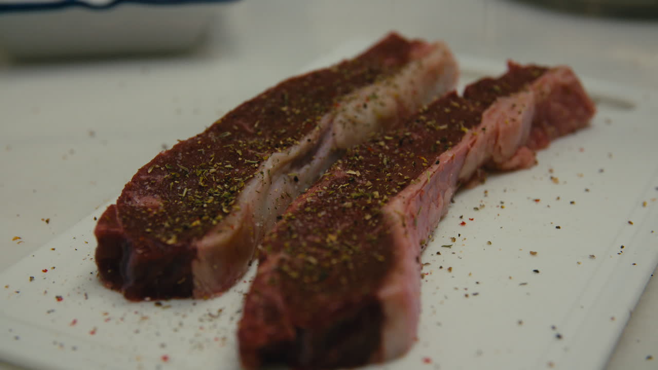 Extreme close-up of raw entrecôte steak coated with salt and freshly ground pepper, resting on a white cutting board. The marbling and texture of the meat showing