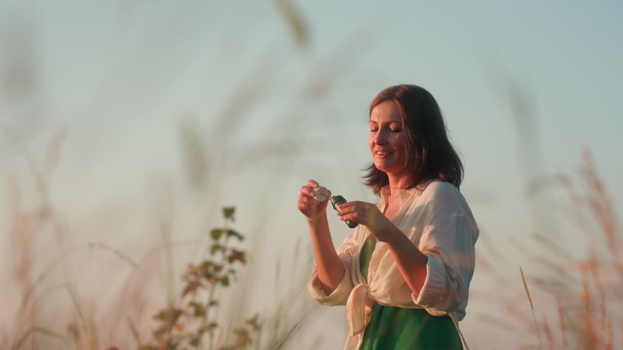Fair skinned woman in white shirt and green dress standing on cliff edge over river valley at sunset, joyfully blowing iridescent soap bubbles drifting among tall grasses in serene nature