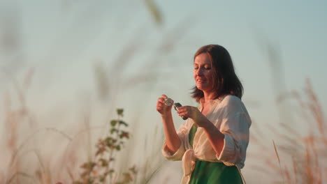 fair skinned woman in white shirt and green dress standing on cliff edge over river valley at sunset, joyfully blowing iridescent soap bubbles drifting among tall grasses in serene nature