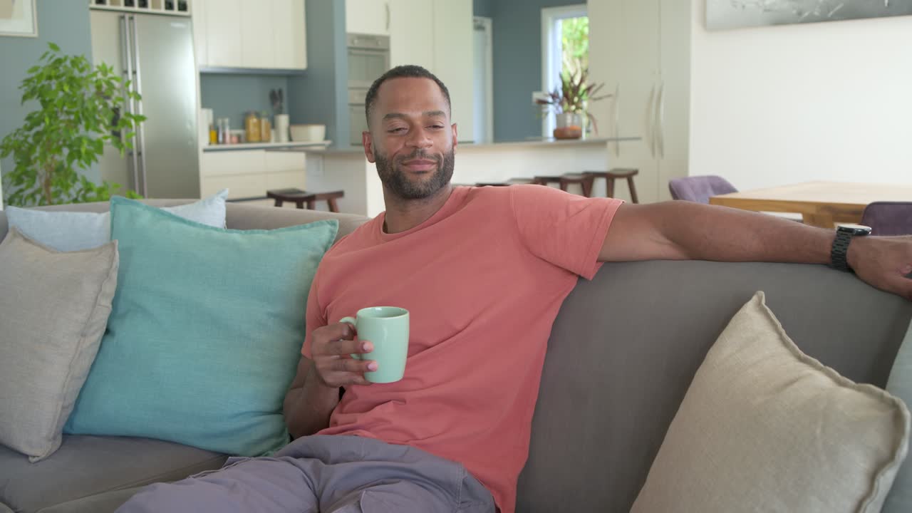 Uomo afroamericano maturo in maglietta color corallo che si rilassa sul divano, sorseggiando da una tazza verde, con la telecamera che si avvicina.