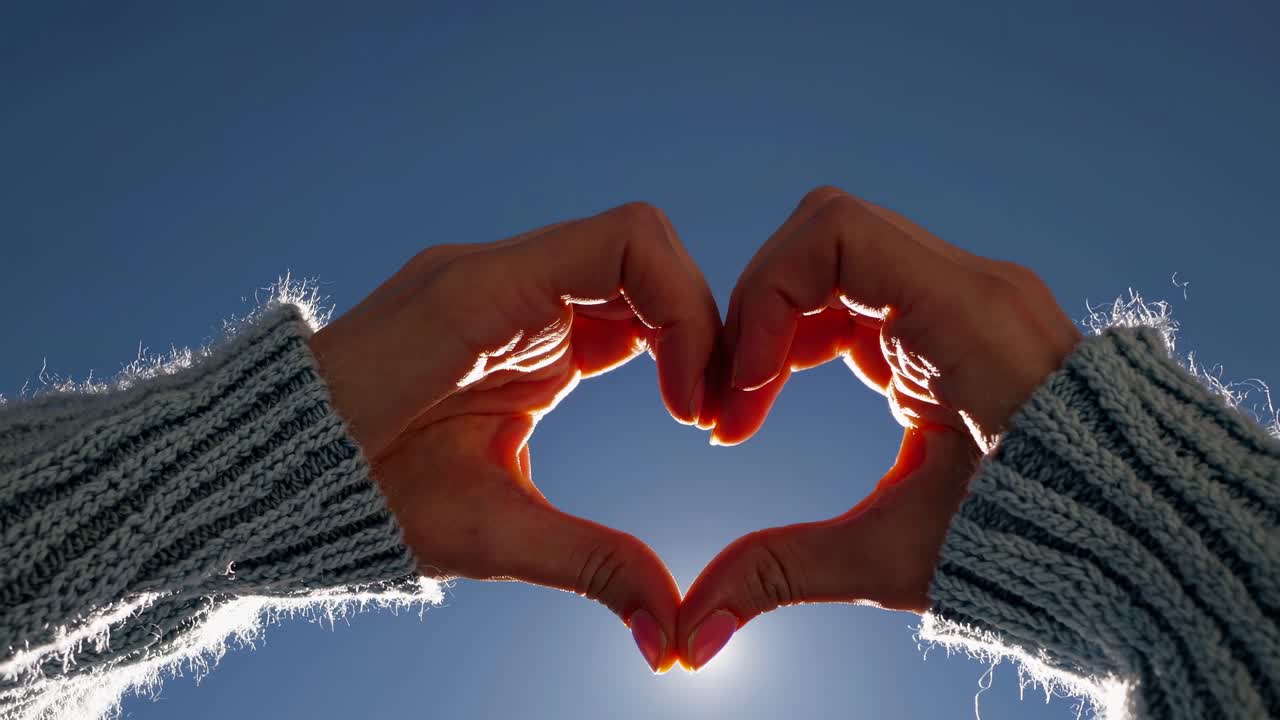 Low-angle video shot of hands forming a heart shape against a clear blue sky, emphasizing warmth