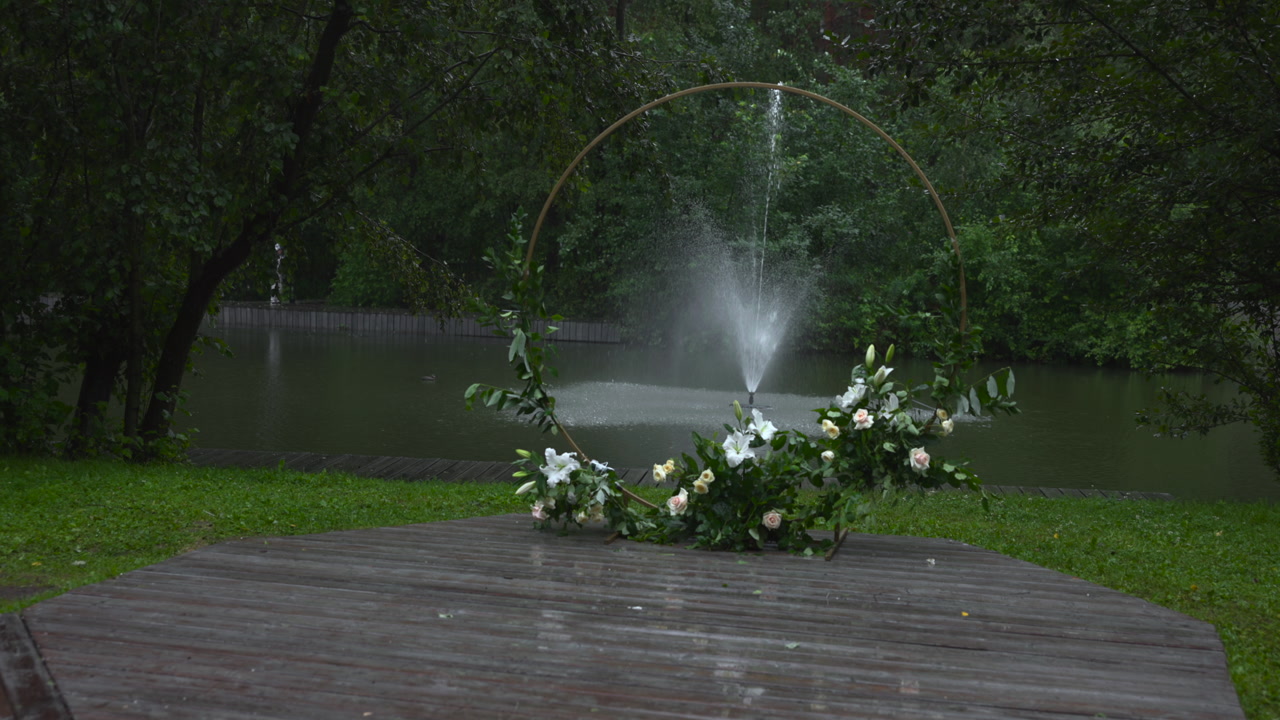 Lakeside Ceremony Arch With Floral Garland, Wooden Platform Wet From Rain, Central Fountain Spray Framed . Media