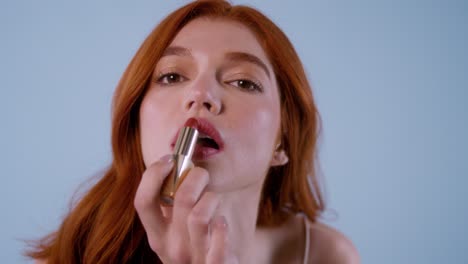 Fair-skinned redhead woman applying lipstick gently, close-up shot showing expression and makeup details.