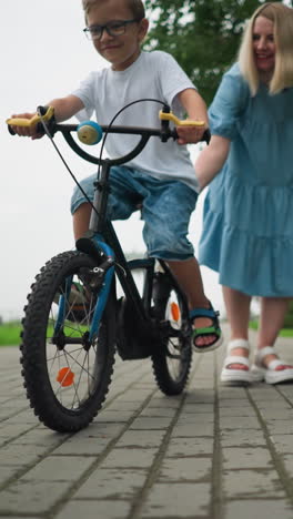 una mujer con una túnica azul ayuda a su hijo a mantener el equilibrio mientras monta su bicicleta por un camino de ladrillo en un parque