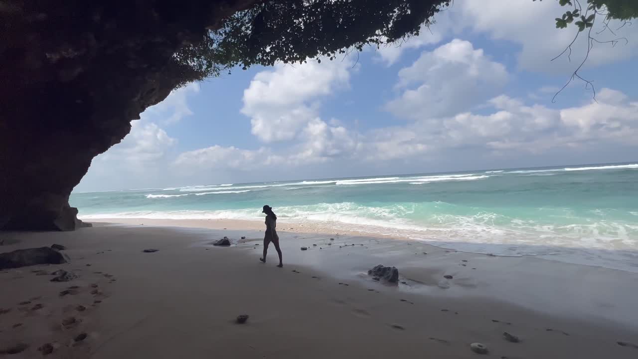 비키니를 입은 젊은 소녀가 은둔한 녹색 그 ⁇  해변 동굴을 산책합니다.