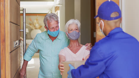 Delivery-man-delivering-package-to-senior-caucasian-couple-wearing-face-masks-at-home