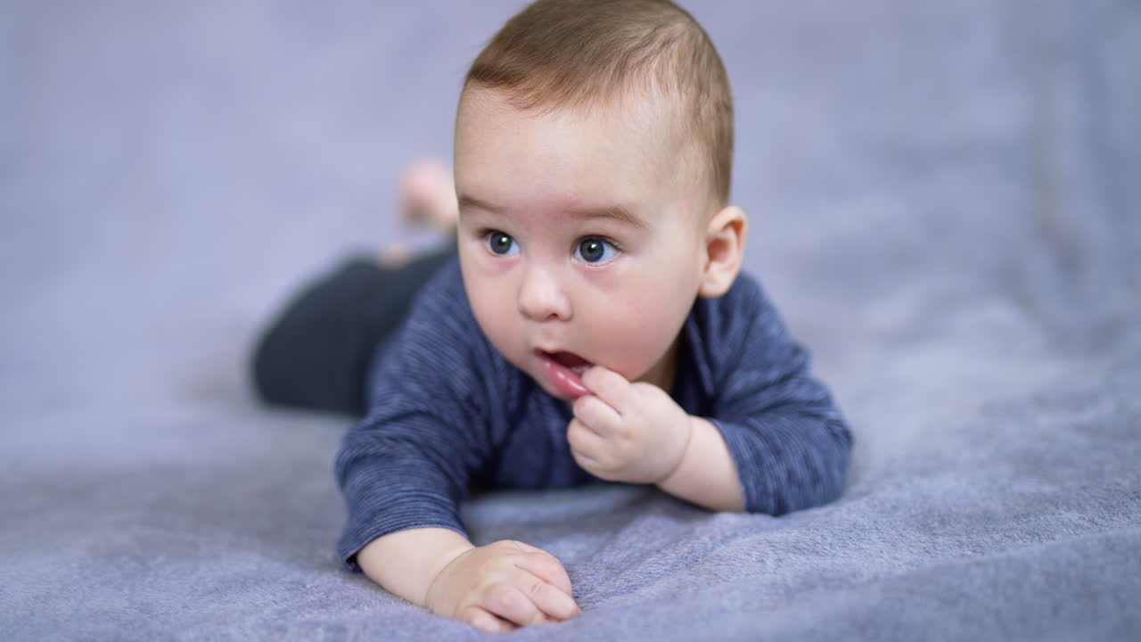 Adorable baby boy pulls his lower lip while lies on bed. Sweet little toddler chews his finger and looks around. Grey blurred backdrop.