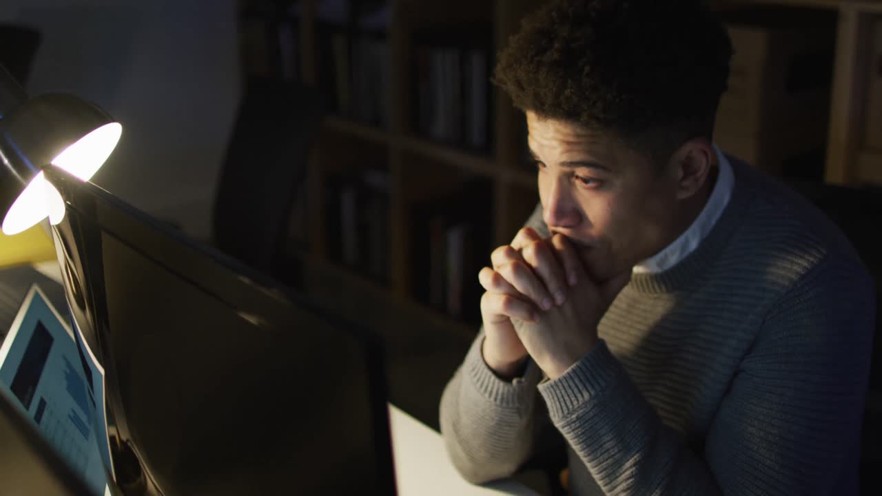 Vídeo de homem birracial cansado usando um computador, trabalhando até tarde no escritório.