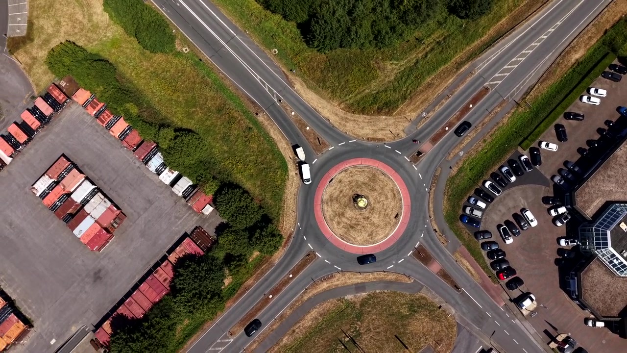 Cars navigate circular roundabout intersection in USA, encircled by industrial shipping containers, busy parking lots and green tree-lined roadside embankments below. Aerial drone top down circling