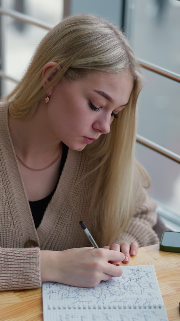 Young lady seated thoughtfully writing with hand on jaw, engaged in deep thought, with coffee cup on table and background showing busy urban setting in winter