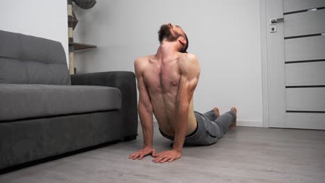 Young-bearded-man-without-t-shirt-doing-yoga-cobra-pose-at-home-in-his-living-room
