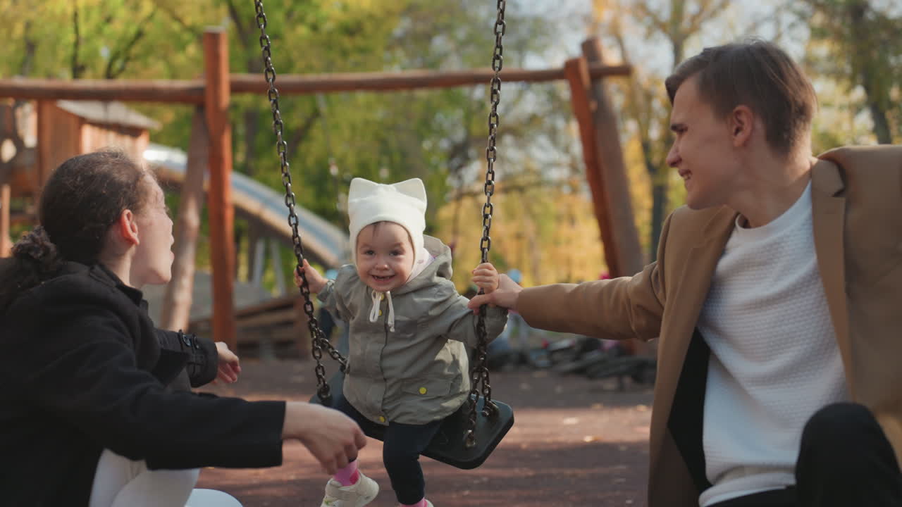 Genitori caucasici bianchi che acclamano felicemente il bambino, bambino con il berretto che ride sotto gli alberi autunnali al parco giochi, filmato caldo e spontaneo che mostra la gioia della coppia sposata, l'unione e la famiglia affettuosa