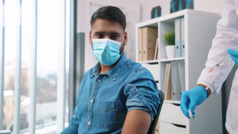 Retrato-De-Primer-Plano-De-Un-Médico-Adulto-Experimentado-De-Mediana-Edad-Que-Hace-Una-Vacuna-Covid-A-Un-Joven-Paciente-Hindú-Con-Mascarilla-Médica-Sentado-En-El-Hospital,-Vacunación-Contra-El-Coronavirus,-Tratamiento-Contra-El-Virus