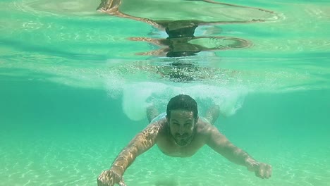 Hombre-Buceando-En-Agua-De-Mar-Azul-Poco-Profunda-Y-Nadando-Bajo-El-Agua-Hacia-La-Cámara-Sonriendo---Mostrando-Los-Pulgares-Hacia-Arriba