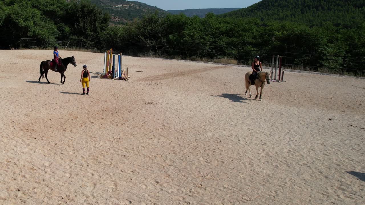 Slow approach drone shot moves toward a horse trainer observing pupils as they ride the arena