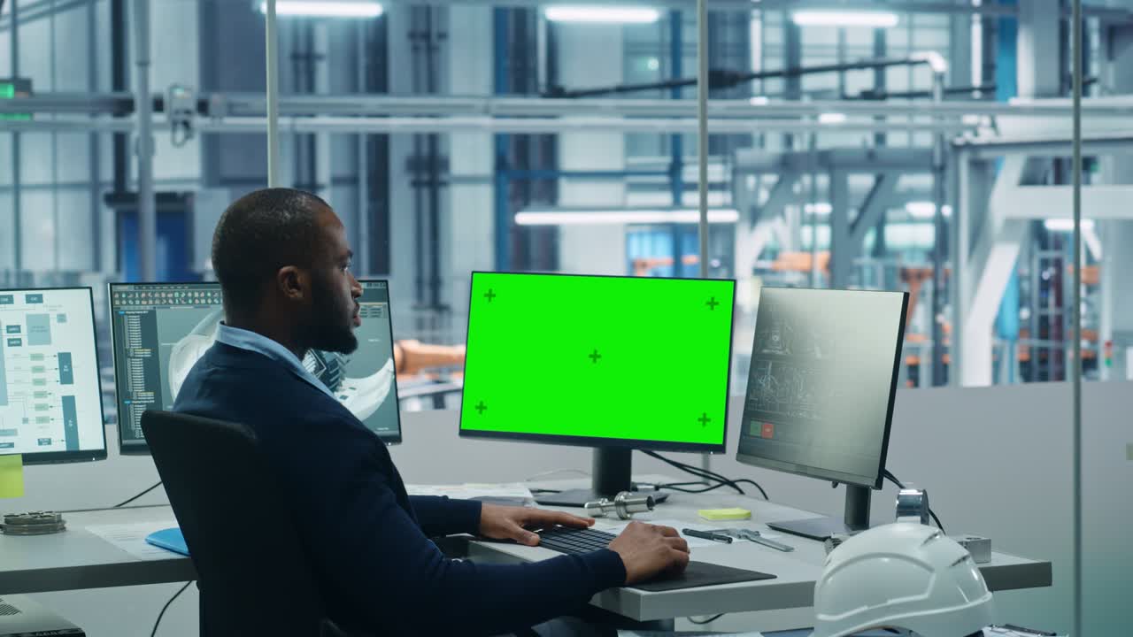 Car Factory Office: Black Engineer Works on a Computer, Two Monitor Screens Show Chroma Key Green Screen and Monitoring of Automated Robot Arm Assembly Line Manufacturing High-Tech Electric Vehicles