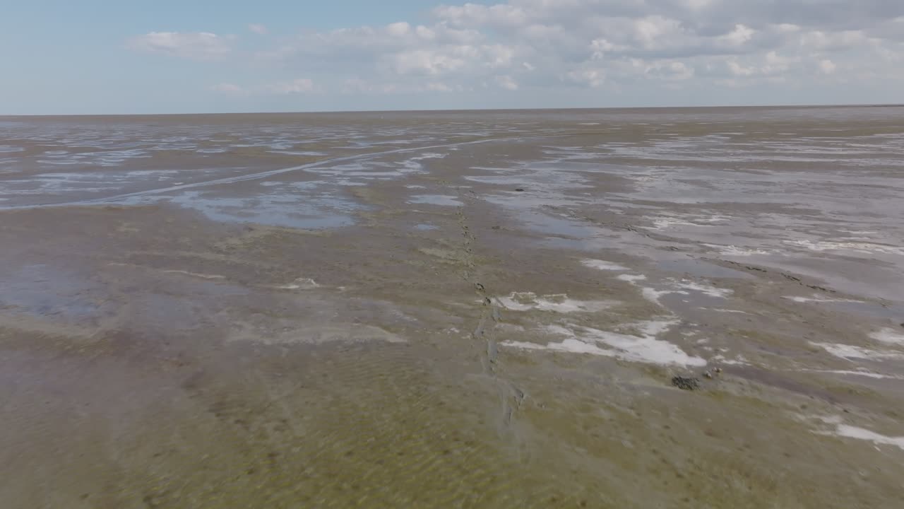 Wide aerial sweep across expansive muddy tidal flats with wet sheen and irregular patterns at low tide; no vessels in frame. Wadden Sea