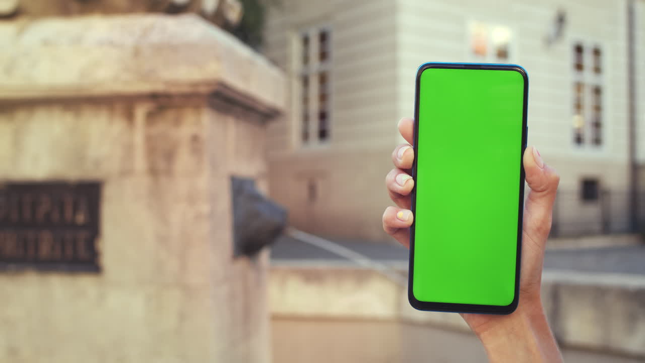 A hand holding a smartphone with a green screen outdoors